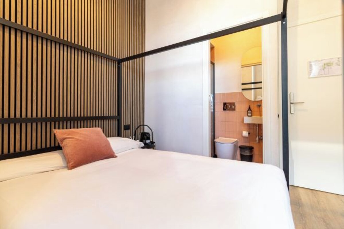 A modern hotel room with a bed, a nightstand, a wall-mounted heater, and a door leading to a bathroom.