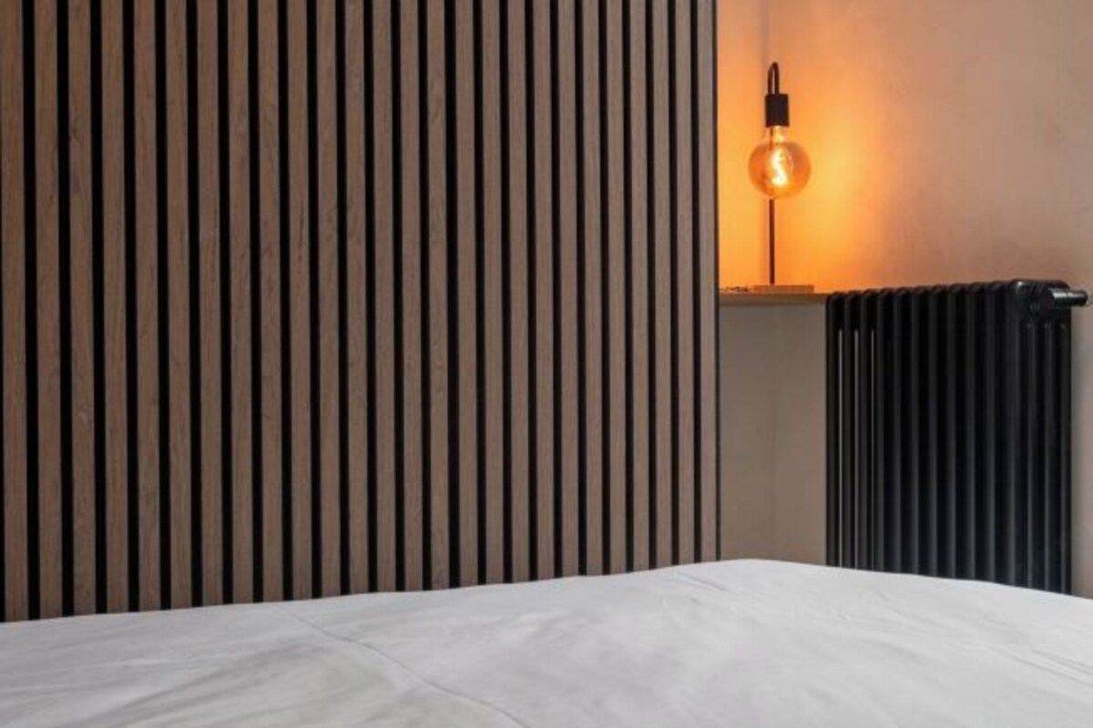 A room with a bed, a vertical wooden panel wall, a wall-mounted light, and a radiator.