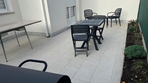 Een terras met een tafel, stoelen, een raam en een bloembed met groene planten.