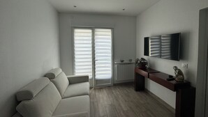 Een woonkamer met een bank, een aan de muur bevestigde televisie en een raam met jaloezieën.