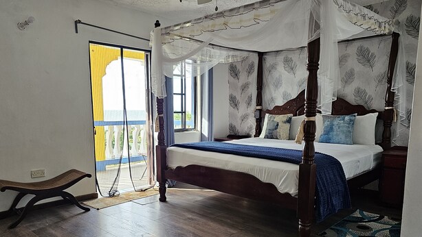 Una cama con dosel y cuatro columnas con estructura de madera, un edredón azul y ropa de cama blanca. A la izquierda de la cama hay un pequeño banco de madera, y una alfombra estampada en el suelo.