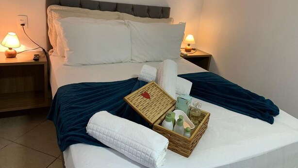 A neatly made bed with a blue blanket, white pillows, and a basket of toiletries on a white bedspread. - Pousada Perola da Praia (Penha)