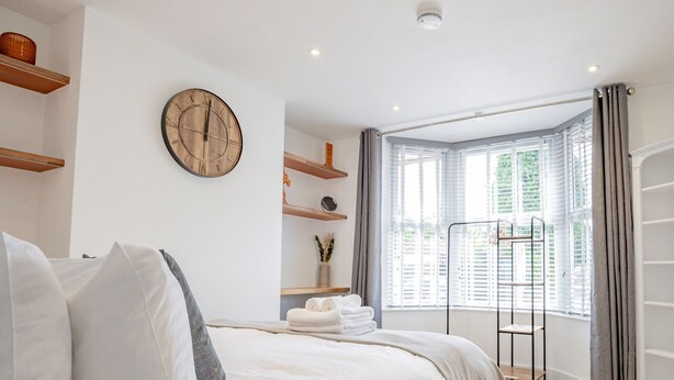 A modern bedroom with a large bed, a wooden clock, built-in shelves, a window with blinds, and a walk-in closet.
