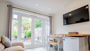 A modern living room with a sofa, a dining table with chairs, a flat-screen TV mounted on the wall, and a sliding glass door leading to a garden.