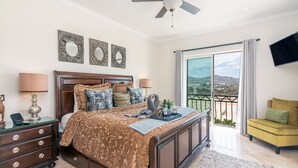 A bedroom with a large bed, a nightstand, a dresser, a balcony with a view, and a TV mounted on the wall.