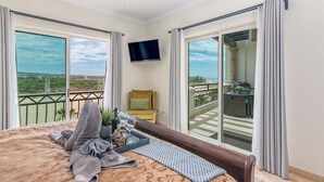 A hotel room with a large bed, a TV, and a balcony with a view of the city and beach.