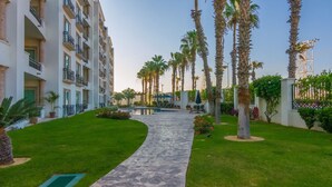A pathway leading to a pool surrounded by palm trees and a multi-story building with balconies.