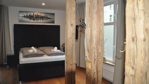Une chambre avec un grand lit, une œuvre d’art représentant un paysage urbain accrochée au mur et une vue sur l’extérieur par une fenêtre.