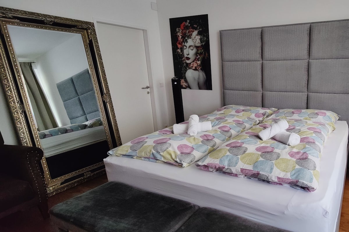 A neatly made bed with a colorful patterned duvet, a large mirror with an ornate frame, a modern headboard, a bedside lamp, and a small bedside table.