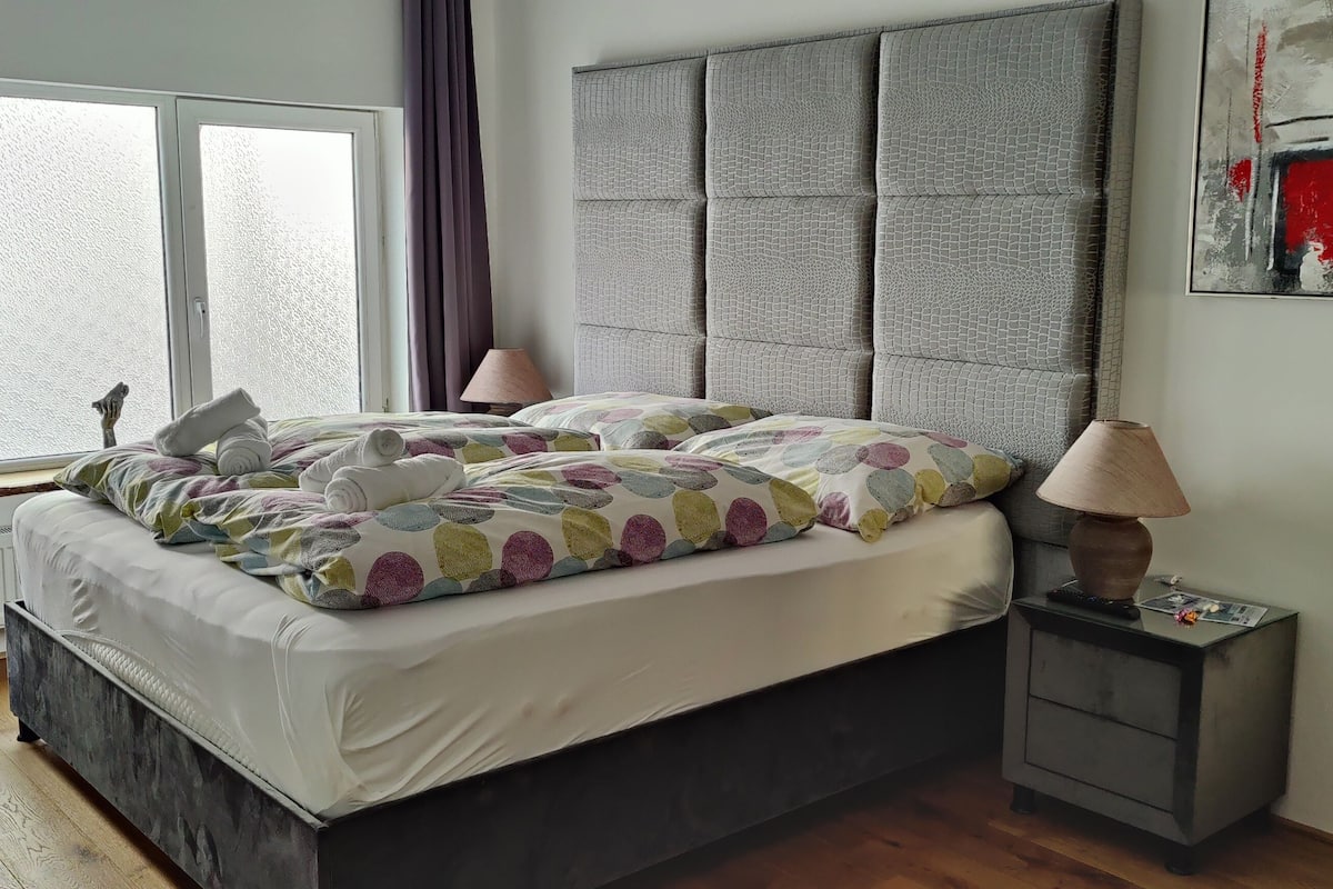A bedroom with a large bed, a headboard, a bedside table with a lamp, and a window with curtains.