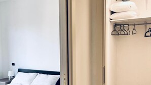 A hotel room with a bed, a wooden nightstand, a wardrobe, and a closet with hanging clothes.