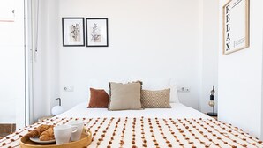 A neatly arranged bedroom with a bed, pillows, a tray with croissants and coffee cups, and two framed botanical prints on the wall.