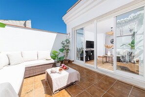 Un patio iluminado por el sol con un sofá blanco, una mesita con un sombrero y un cuenco, y una planta en maceta.