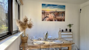 A dining area with a wooden table, clear plastic chairs, a vase with pampas grass, a wine bottle, and a painting of a sunset over a beach. - Appartement à Bandol Avec Piscine et Accès mer (Bandol)
