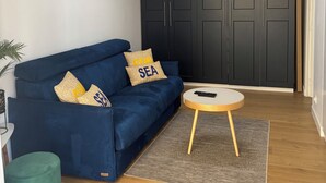 A living room with a blue sofa, a round coffee table, a dark wardrobe, and a painting of a sailboat. - Appartement à Bandol Avec Piscine et Accès mer (Bandol)