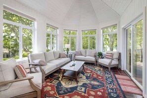 Une véranda aux murs blancs, dotée de grandes fenêtres, d’un tapis à motifs et offrant différents types de sièges, notamment un canapé et des fauteuils.