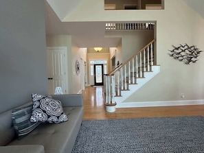 Ein geräumiges Wohnzimmer mit grauem Sofa, gemusterten Kissen und einem grauen Teppich. Eine Holztreppe mit weißen Spindeln führt zu einer Tür am Ende des Flurs.
