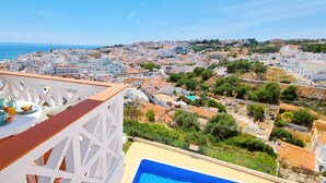 Un balcon avec une table préparée pour le petit-déjeuner, donnant sur une ville côtière avec une piscine.