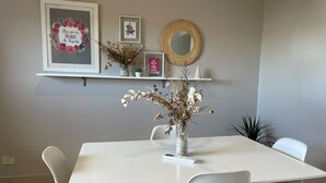 Un coin repas avec une table et des chaises blanches, un vase contenant des fleurs séchées et des éléments de décoration murale, notamment une citation encadrée et un miroir.