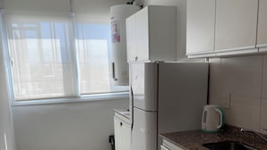 Une cuisine avec des meubles blancs, un réfrigérateur en acier inoxydable, un plan de travail en granit et une fenêtre avec des stores.