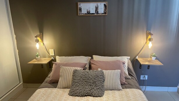 Un lit bien fait, avec une literie à motifs, deux tables de chevet avec des lampes, un tableau encadré au mur et une petite plante sur chaque table.