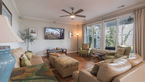 A living room with a beige sofa, a patterned ottoman, a coffee table, and a ceiling fan. There are two windows with curtains, a floor lamp, and framed pictures on the wall. - Creek-Side Bliss: Unwind and Create Lasting Memories at Our Beautiful Retreat (Pasadena)