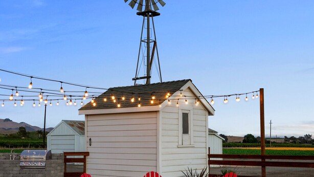 Ein kleines, weißes Gebäude mit Windmühle, umgeben von roten Adirondack-Stühlen und einer Feuerstelle, mit Lichterketten darüber.