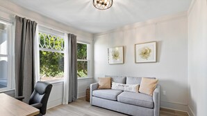 Ein modernes Wohnzimmer mit einem grauen Sofa, einem Holzesstisch und einem schwarzen Bürostuhl. Zwei Fenster mit Vorhängen sowie zwei gerahmte Blumenbilder an der Wand.