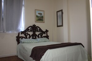 A bedroom with a wooden headboard, a bed with a white sheet and a patterned pillow, a brown throw blanket, and two framed pictures on the wall. - Anfitrioca Aconchegante Apartamento 10 Minutos Metrô da Glória (Glória Rio de Janeiro)