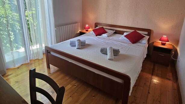 Ein ordentlich bezogenes Bett mit weißen Leinen und roten Kissen, ein Holzboden und ein Fenster mit durchscheinenden Vorhängen.