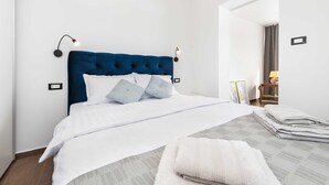 Una cama bien tendida con cabecera capitonada, ropa de cama blanca y algunos almohadones. Se observa una mesita de noche con una lámpara y un cuadro enmarcado. La habitación tiene paredes blancas y suelo de madera.