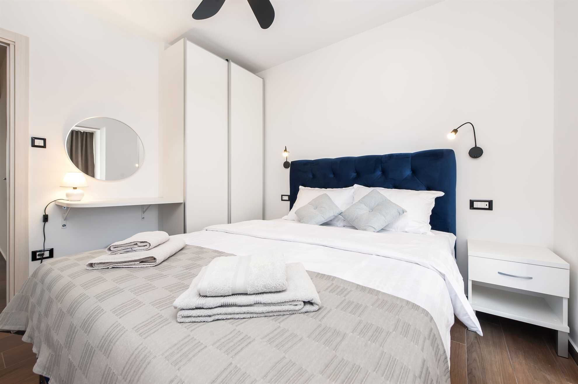 A modern hotel room with a large bed, a bedside table, a nightstand, a round mirror, and a wall-mounted light fixture.