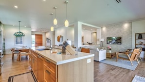 A modern kitchen with wooden cabinets, a central island, pendant lighting, and a dining area with chairs.