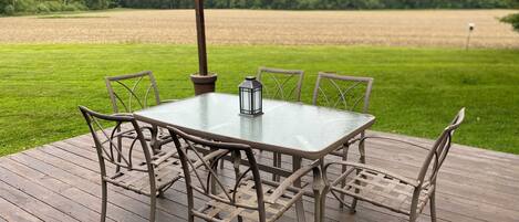 A wooden deck with a glass table and chairs, surrounded by a grassy field and trees.