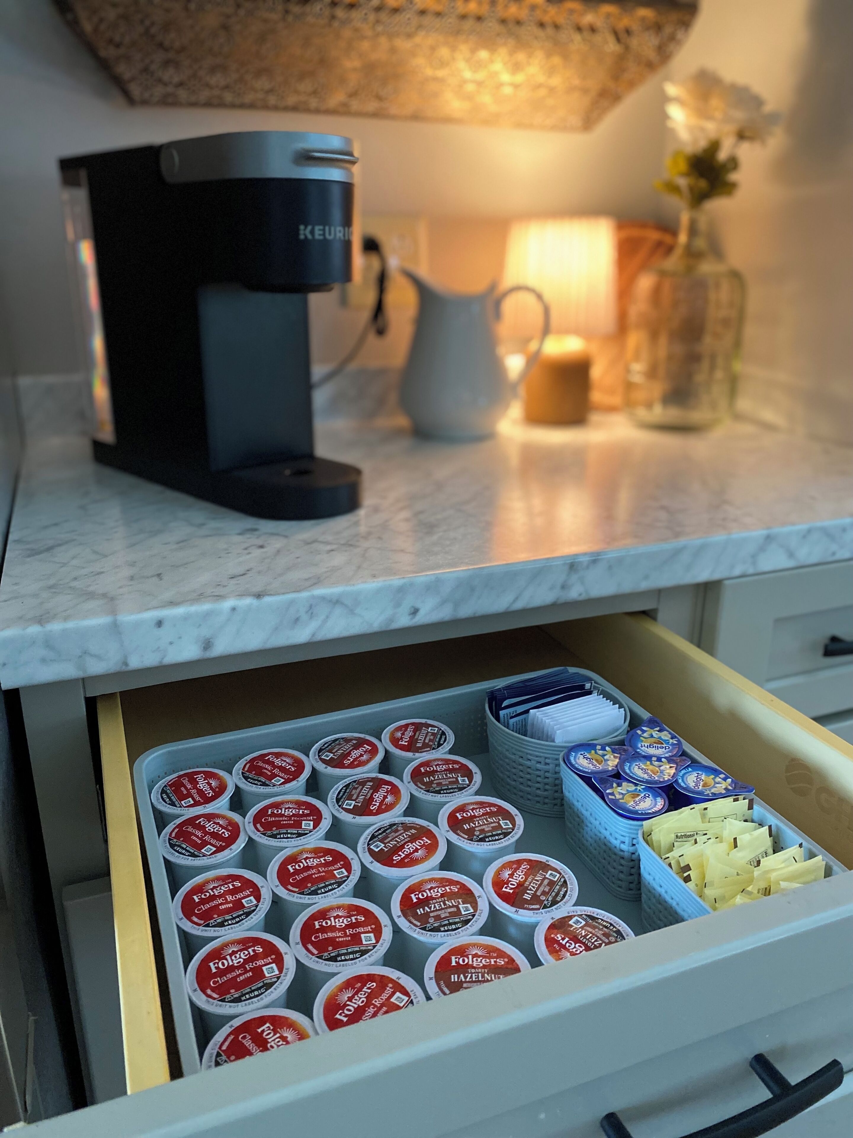 Eine Küchenarbeitsplatte mit einer Kaffeemaschine, einer weißen Kanne und einer Vase mit Blumen. Darunter befindet sich eine Schublade mit Kaffeekapseln und ein Serviettenkorb.