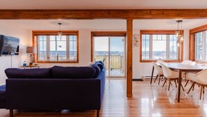 A spacious living room with a dark blue sofa, wooden flooring, large windows, a dining area with white chairs, and a TV mounted on the wall.