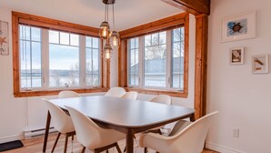 A dining area with a large table and chairs, wooden beams, large windows, and hanging light fixtures.