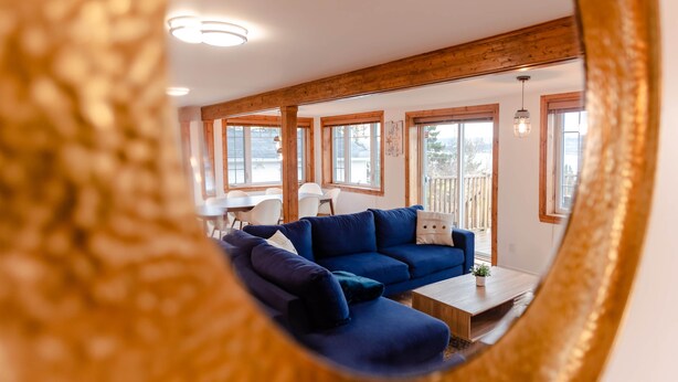 A cozy living room with a blue sofa, wooden beams, large windows, and a dining area with white chairs.