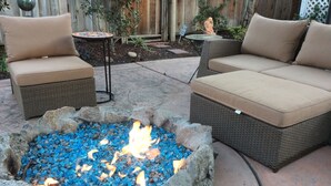 Une terrasse extérieure avec deux canapés en osier dotés de coussins beige, une petite table ronde et un foyer agrémenté de pierres bleues.