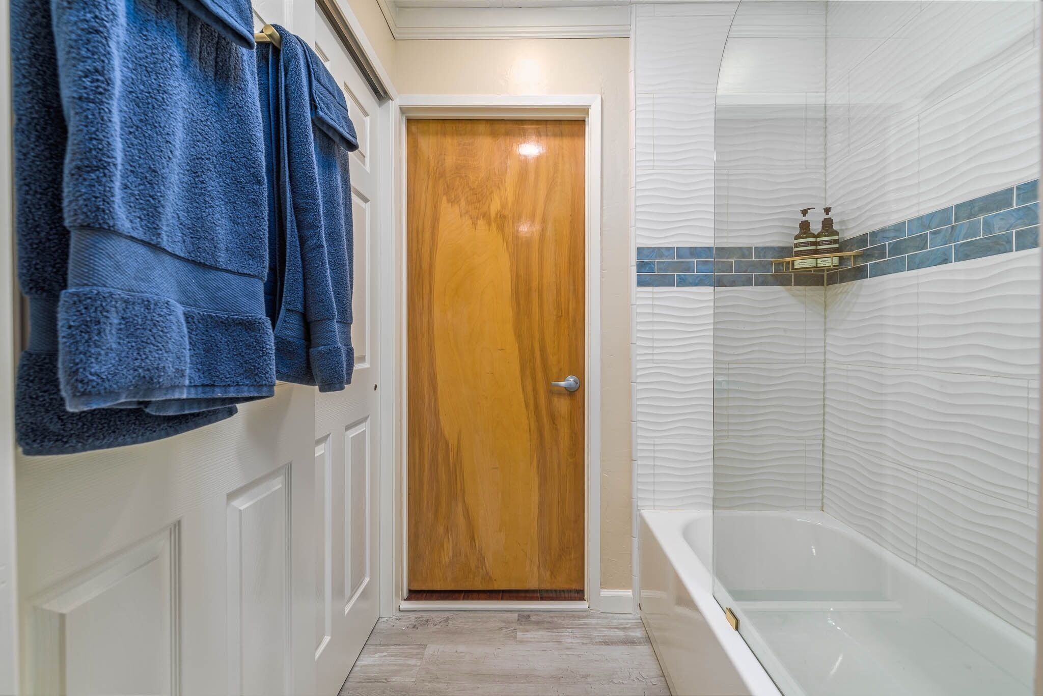 Un baño con puerta de madera, ducha con diseño de azulejos ondulados y un perchero con toallas azules.