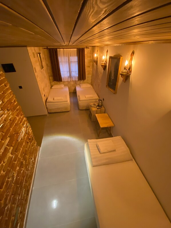 A narrow, dimly lit room with two beds, a small table, and wall-mounted lights.