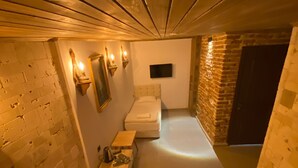 A narrow, dimly lit room with two beds, a small table, and a wall-mounted TV.
