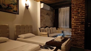A room with two beds, a sofa, and a small table with towels. There is a painting on the wall and a window with curtains.