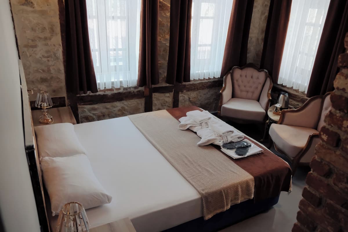 A hotel room with a large bed, two armchairs, a small table with a kettle, and a window with curtains.