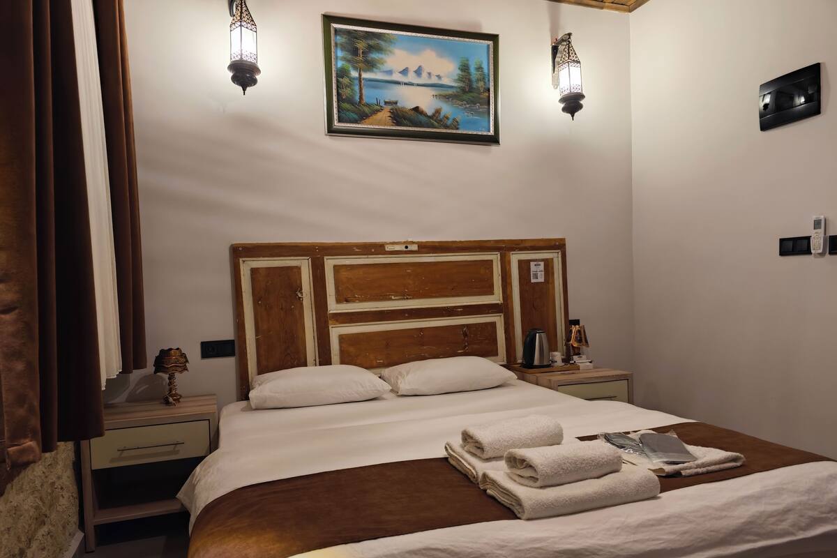 A neatly made bed with white linens, a wooden headboard, and a painting above it. There are two bedside tables with lamps and a small clock.