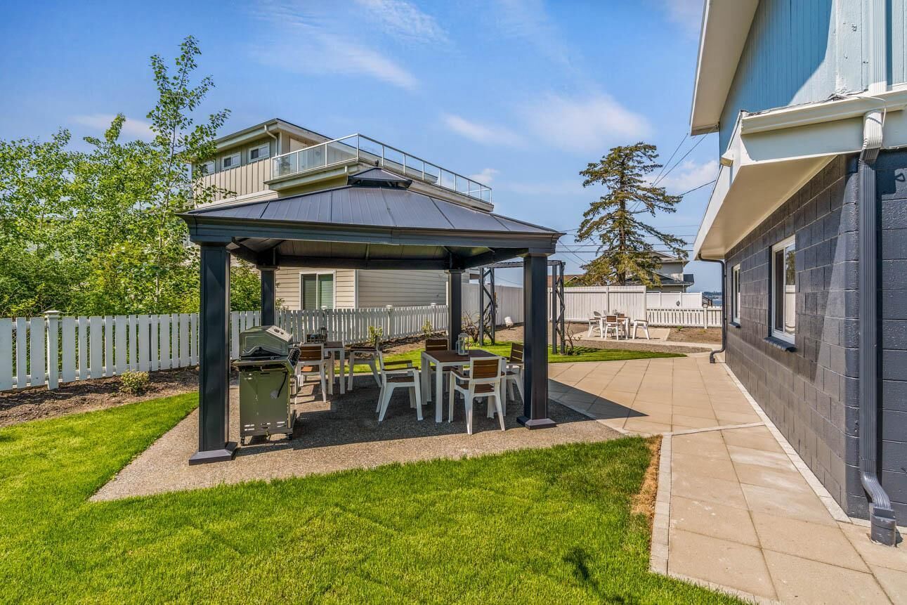 En täckt uteplats med utemöbler och en grill, omgiven av en inhägnad trädgård och ett hus.