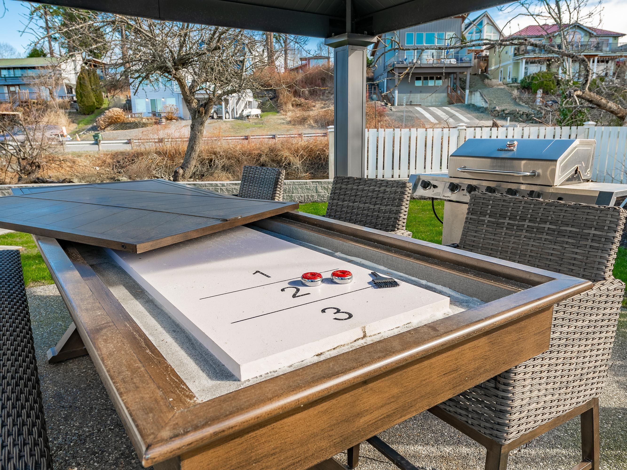 A shuffleboard table with numbered areas, a grill, wicker chairs, and a view of houses in the background.