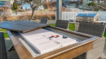A shuffleboard table with numbered areas, a grill, wicker chairs, and a view of houses in the background.