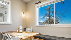 A dining area with a wooden table, white chairs, a bench with a cushion, and a view of a tree through a window.