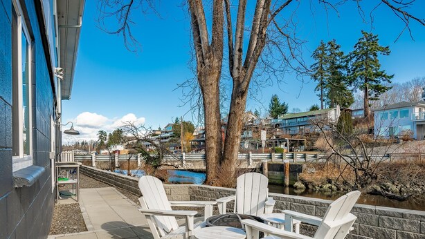 A patio with white Adirondack chairs and a table, overlooking a river and buildings.